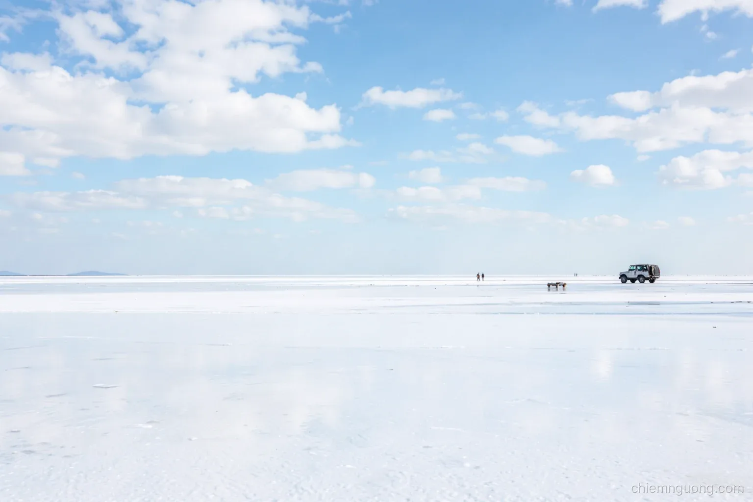 Vast white salt flat covered by a thin layer of water reflecting a sky of scattered clouds, two small distant figures and a parked jeep, horizon line vanishing, pale blue palette