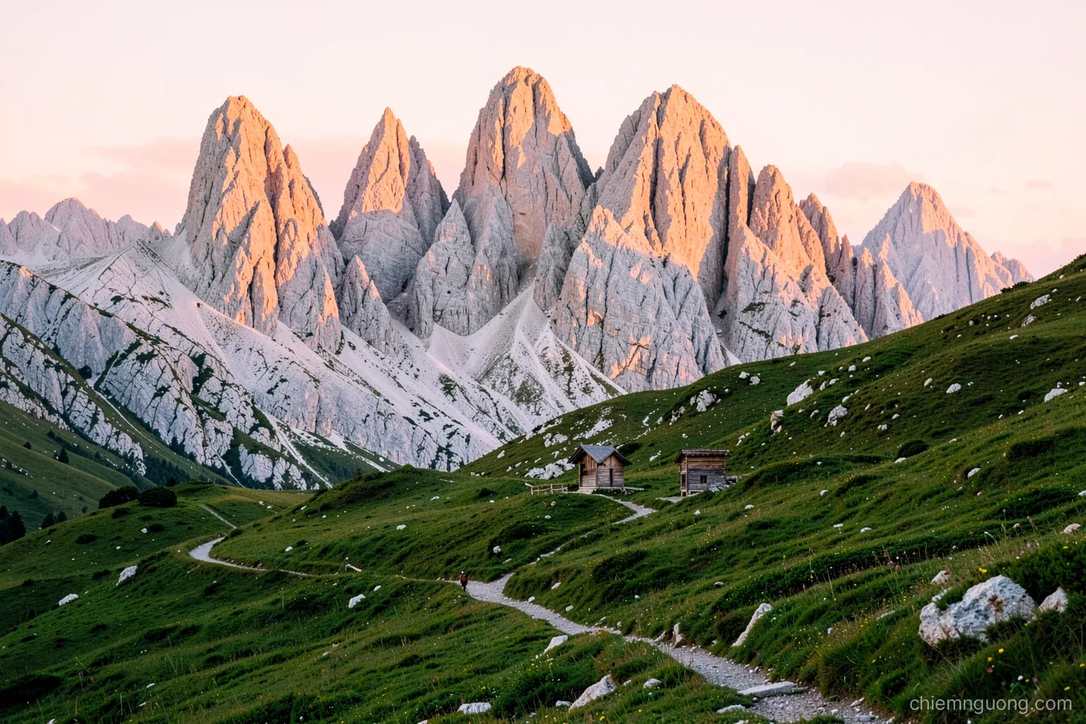 Golden hour over jagged pale limestone peaks glowing pink above a green alpine meadow with a small wooden hut, a hiker small on a winding trail, warm rose and green palette