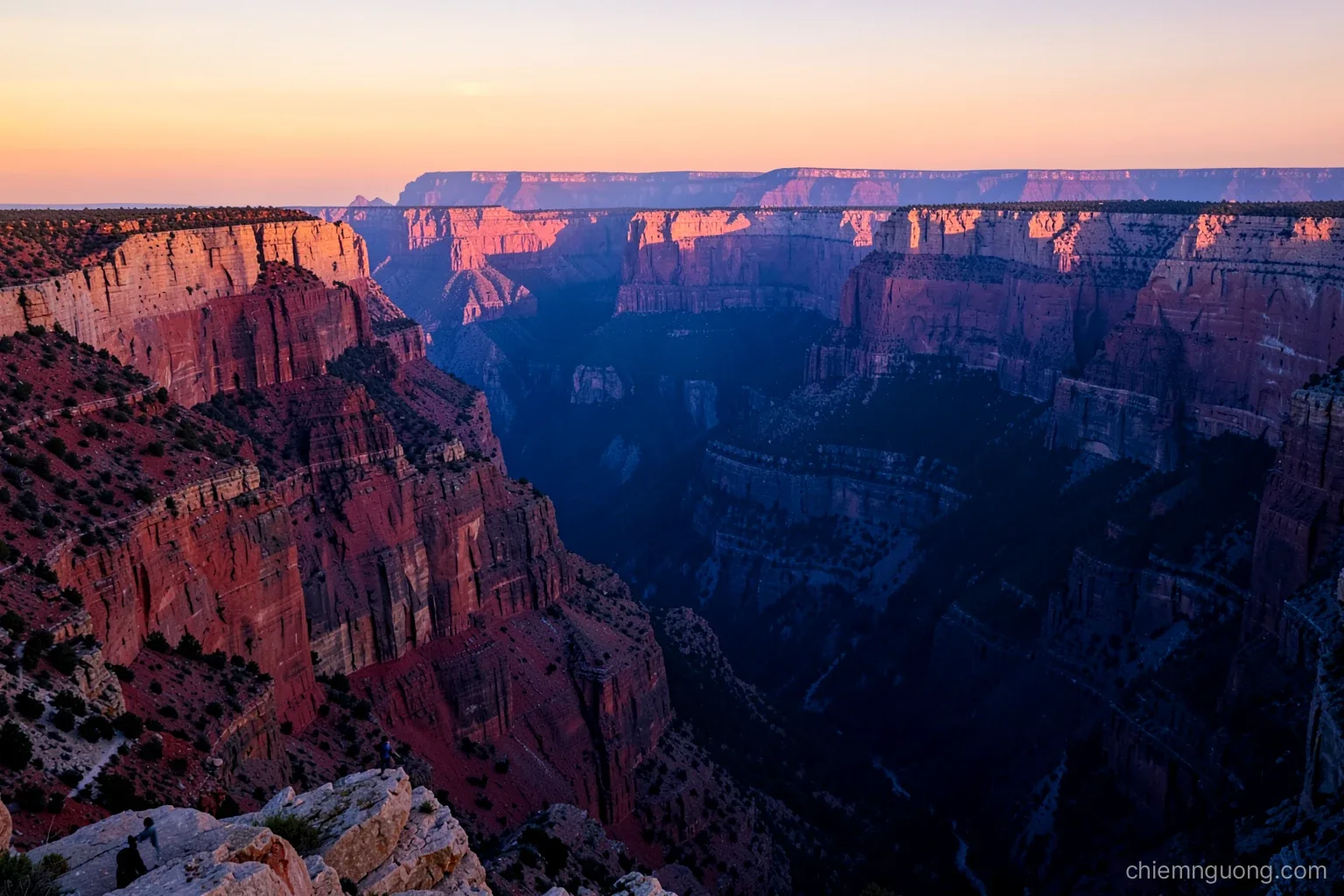 Wide view from a canyon rim at sunset, layered red and cream cliffs stepping down into deep blue shadow, a tiny hiker silhouetted on a rock ledge, warm amber and violet palette