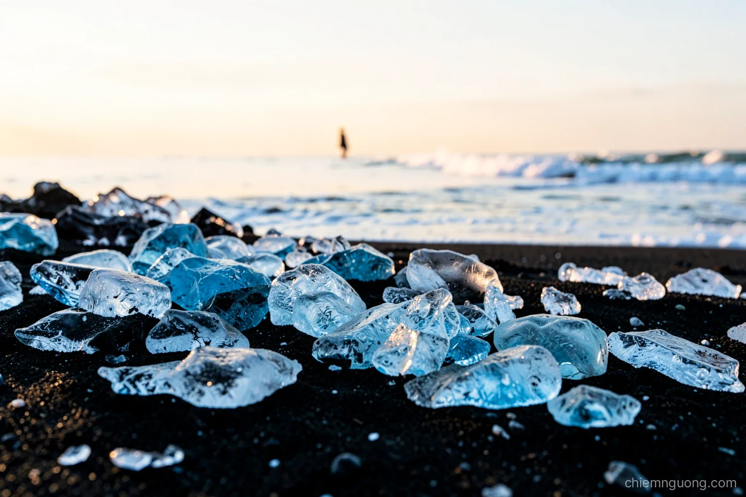 Chunks of clear and blue ice scattered on a black volcanic sand beach at sunrise, gentle surf foaming around them, a distant small figure, low golden light, black white and blue palette