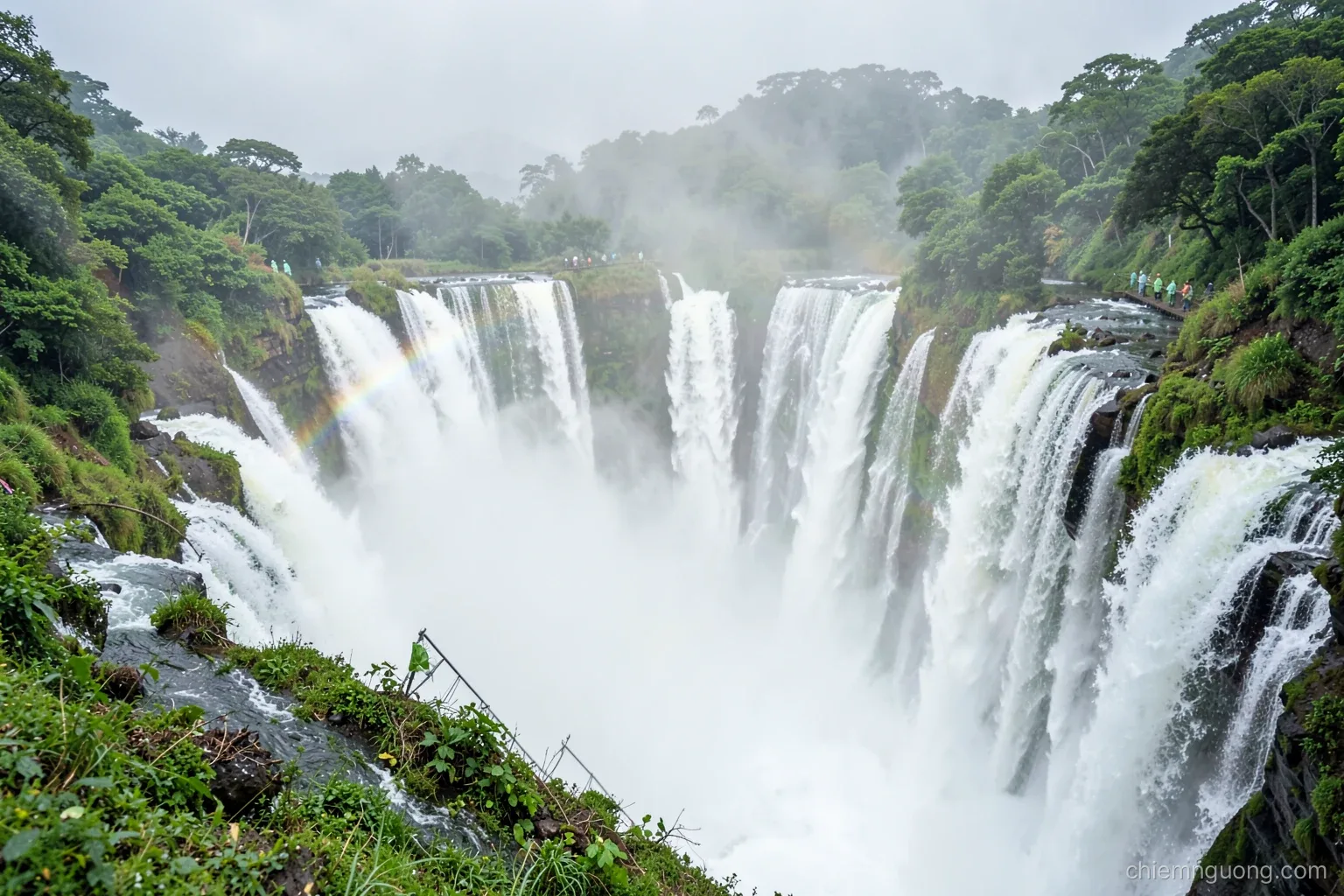 Close wide view of a horseshoe-shaped wall of waterfalls plunging into white mist, a metal walkway with small figures in rain ponchos, a faint rainbow, lush green and white palette